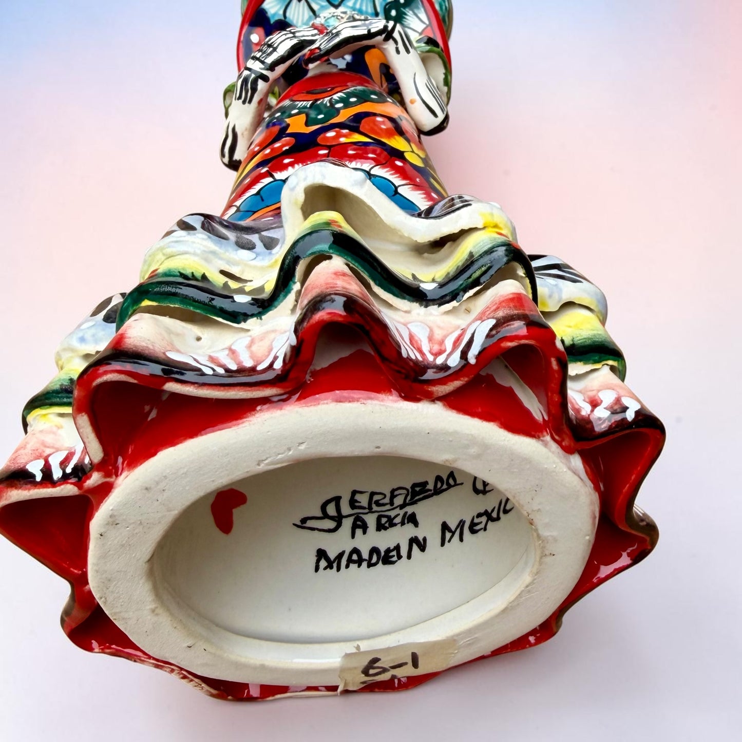 A colorful Day of the Dead talavera skeleton figurine wearing a wide-brimmed red hat and a vibrant floral dress, set against a soft light pink gradient background. bottom showing artist signature.