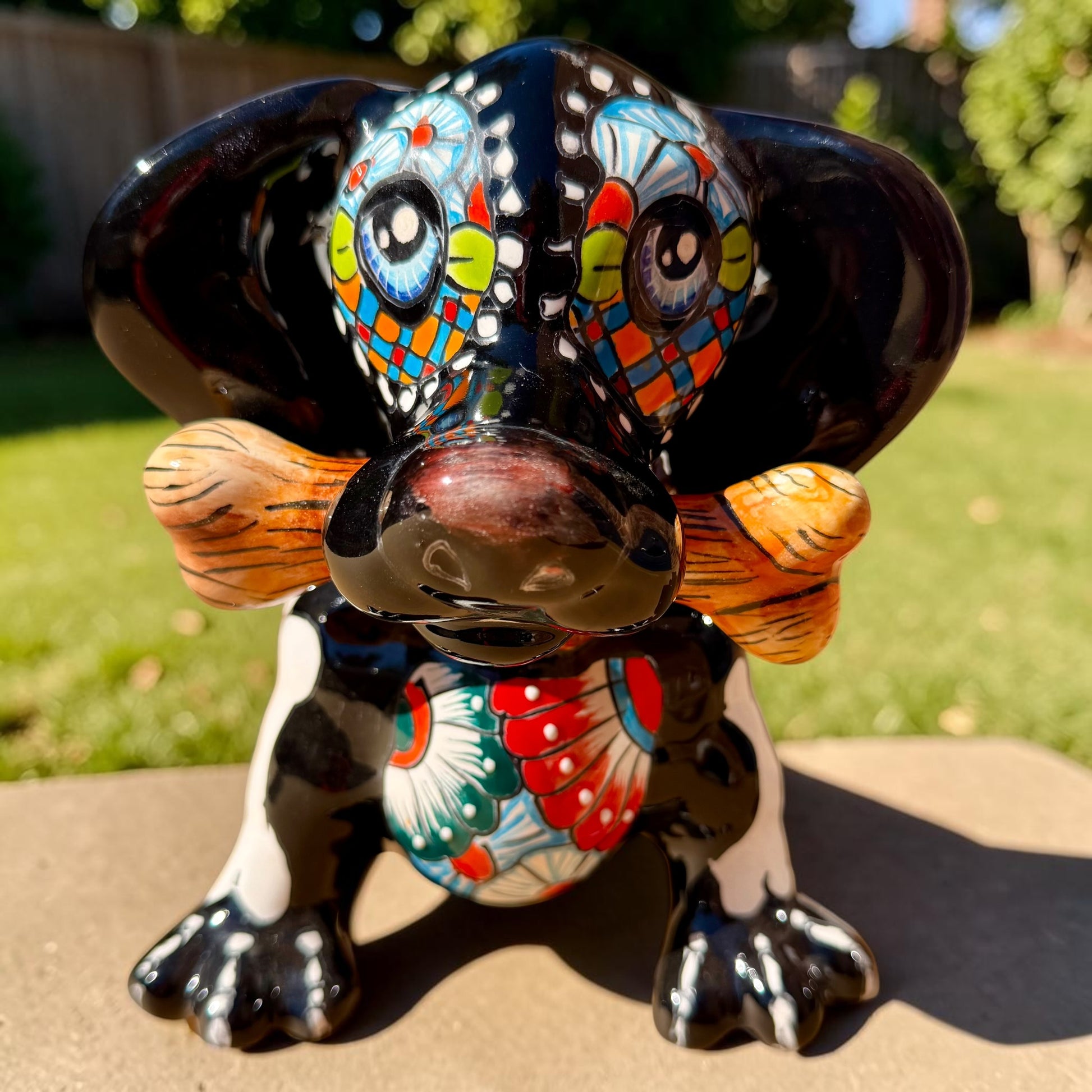 A glossy black talavera day of the dead dachshund figurine decorated with colorful  patterns and hand-painted bones, holding a brown bone in its mouth, set on a green grass lawn. Front view showing bone in mouth and expressive eyes.
