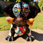A glossy black talavera day of the dead dachshund figurine decorated with colorful  patterns and hand-painted bones, holding a brown bone in its mouth, set on a green grass lawn. Front view showing bone in mouth and expressive eyes.
