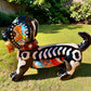 A glossy black talavera day of the dead dachshund figurine decorated with colorful  patterns and hand-painted bones, holding a brown bone in its mouth, set on a green grass lawn. Right side view.