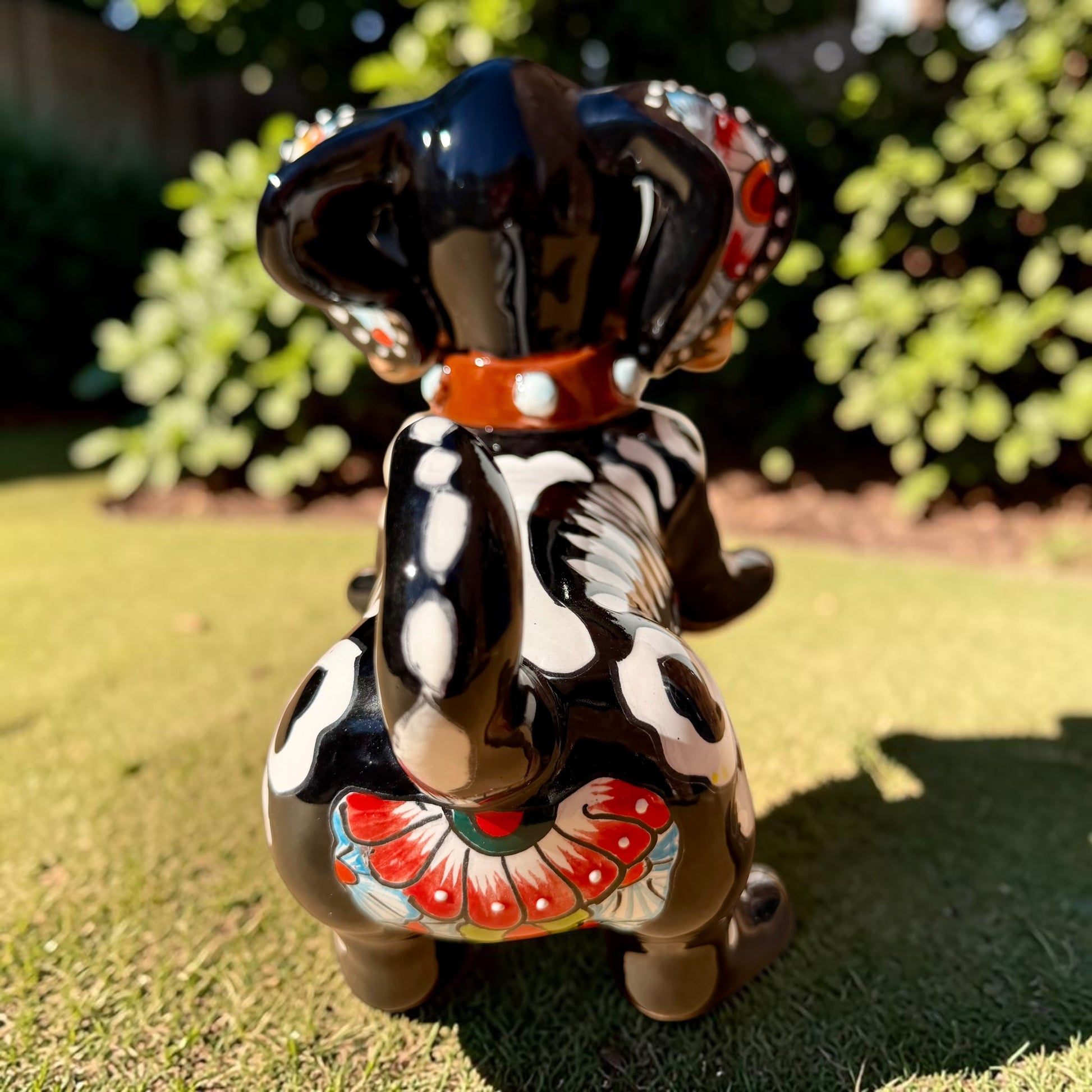 A glossy black talavera day of the dead dachshund figurine decorated with colorful  patterns and hand-painted bones, holding a brown bone in its mouth, set on a green grass lawn. Back view of tail.