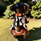 A glossy black talavera day of the dead dachshund figurine decorated with colorful  patterns and hand-painted bones, holding a brown bone in its mouth, set on a green grass lawn. Back view of tail.