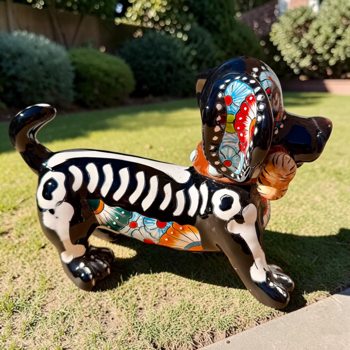 A glossy black talavera day of the dead dachshund figurine decorated with colorful  patterns and hand-painted bones, holding a brown bone in its mouth, set on a green grass lawn. Left side view of puppy.