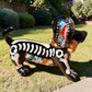 A glossy black talavera day of the dead dachshund figurine decorated with colorful  patterns and hand-painted bones, holding a brown bone in its mouth, set on a green grass lawn. Left side view of puppy.