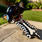 A glossy black talavera day of the dead dachshund figurine decorated with colorful  patterns and hand-painted bones, holding a brown bone in its mouth, set on a green grass lawn. View of the top showing back bonel