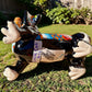 A glossy black talavera day of the dead dachshund figurine decorated with colorful  patterns and hand-painted bones, holding a brown bone in its mouth, set on a green grass lawn. Bottom of dachshund showing artist signature.