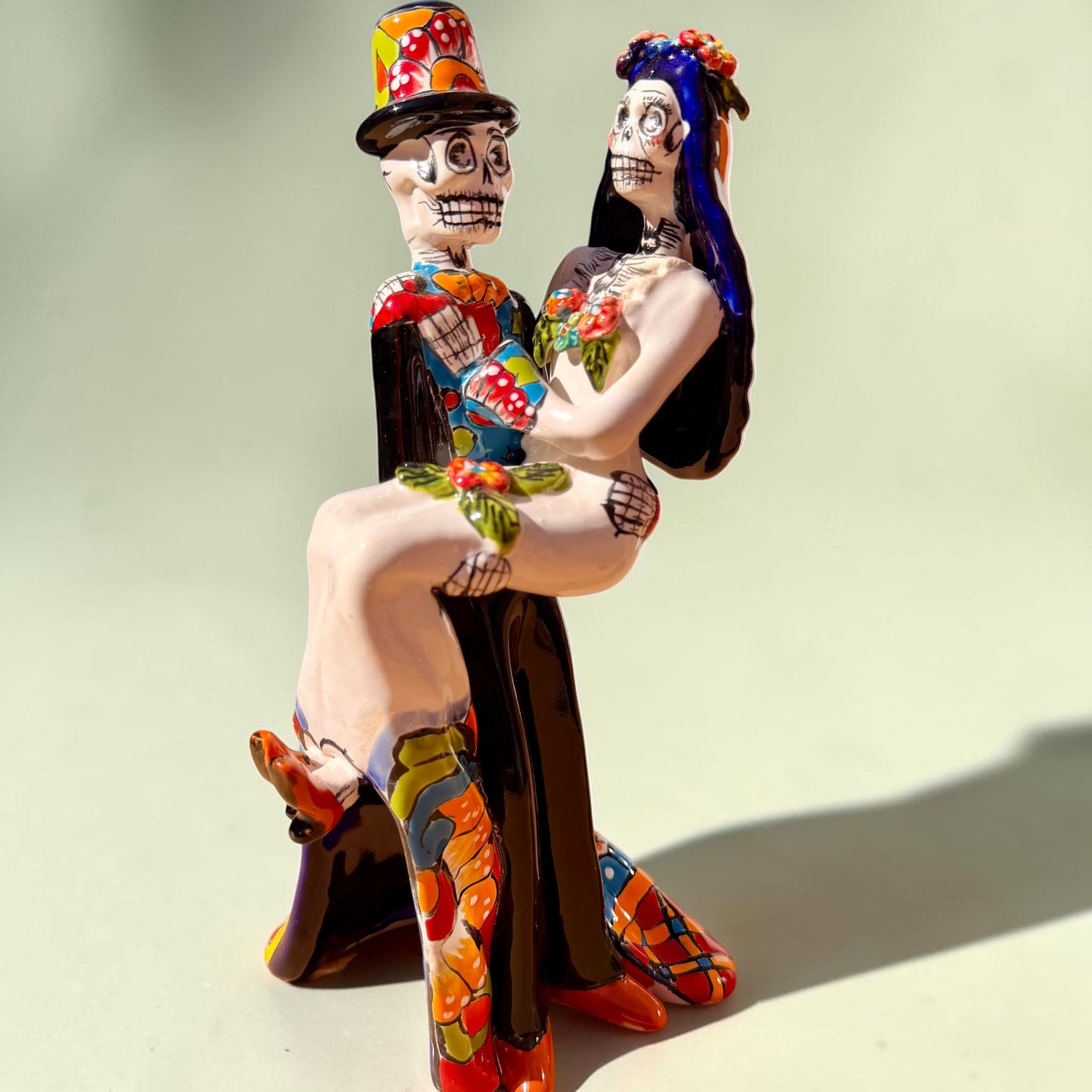 A colorful Day of the Dead figurine of a skeleton bride and groom embracing, with the groom holding the bride. The figurine features vibrant painted details including a orange and white patterned dress, a multicolored hat, and dark hair, set against a plain light background.
