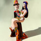 A colorful Day of the Dead figurine of a skeleton bride and groom embracing, with the groom holding the bride. The figurine features vibrant painted details including a orange and white patterned dress, a multicolored hat, and dark hair, set against a plain light background.