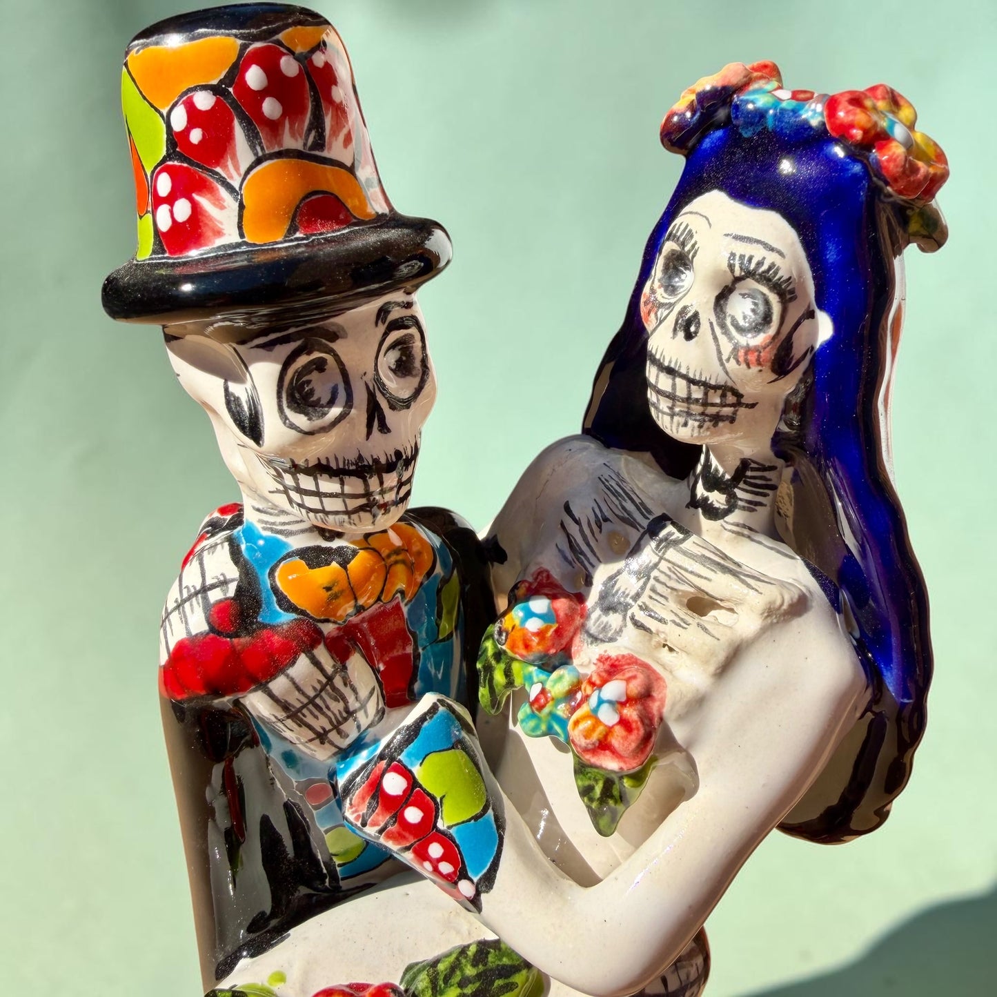 A colorful Day of the Dead figurine of a skeleton bride and groom embracing, with the groom holding the bride. The figurine features vibrant painted details including a orange and white patterned dress, a multicolored hat, and dark hair, set against a plain light background. Close up of faces.
