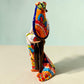 A colorful Day of the Dead figurine of a skeleton bride and groom embracing, with the groom holding the bride. The figurine features vibrant painted details including a orange and white patterned dress, a multicolored hat, and dark hair, set against a plain light background.  Side view of figurine featuring the bride.