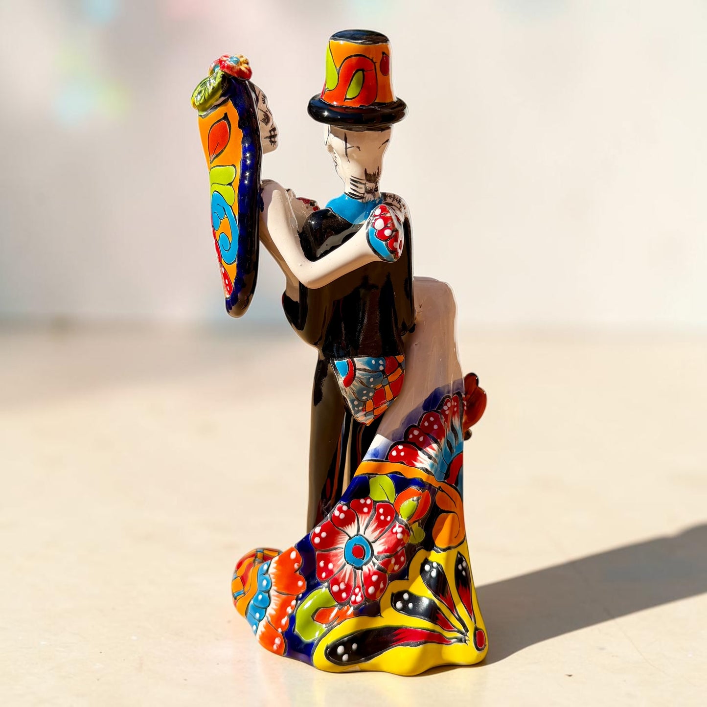 A colorful Day of the Dead figurine of a skeleton bride and groom embracing, with the groom holding the bride. The figurine features vibrant painted details including a orange and white patterned dress, a multicolored hat, and dark hair, set against a plain light background. Back veiw of figurine showing veil and flowing bridal dress.