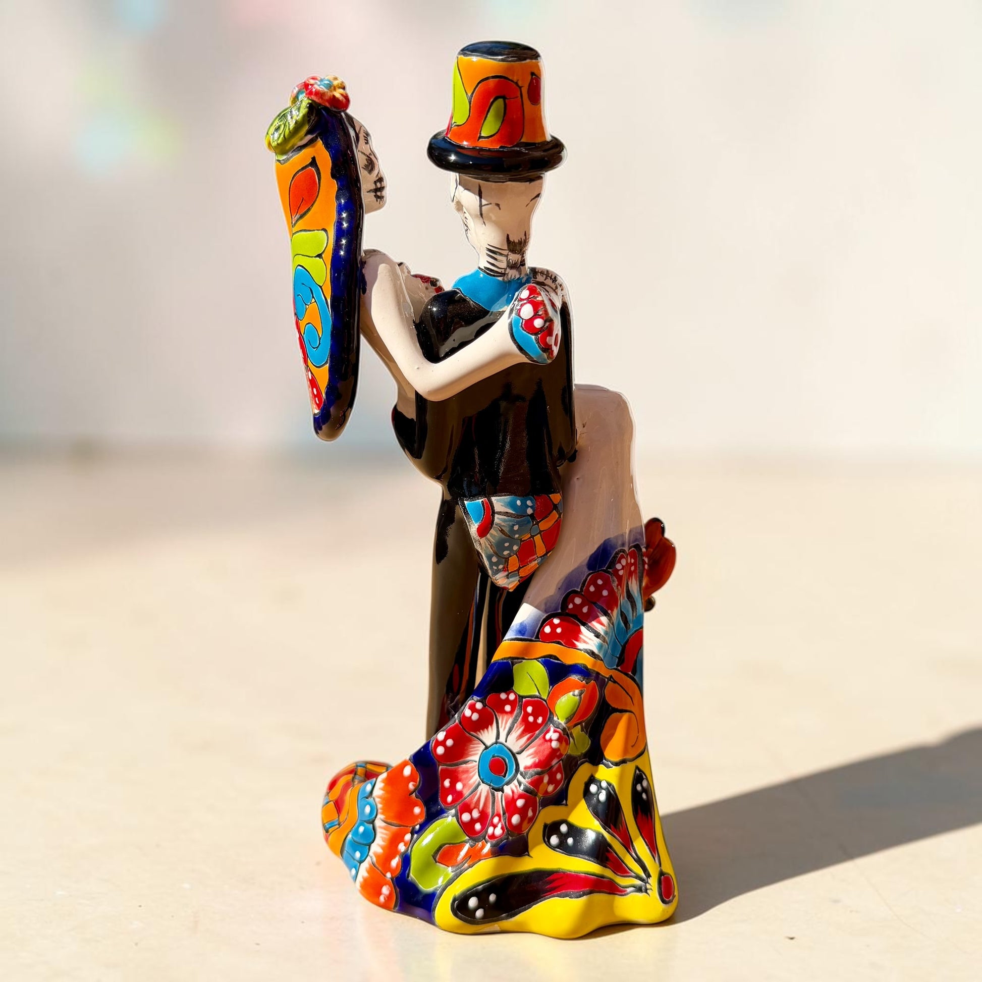 A colorful Day of the Dead figurine of a skeleton bride and groom embracing, with the groom holding the bride. The figurine features vibrant painted details including a orange and white patterned dress, a multicolored hat, and dark hair, set against a plain light background. Back veiw of figurine showing veil and flowing bridal dress.