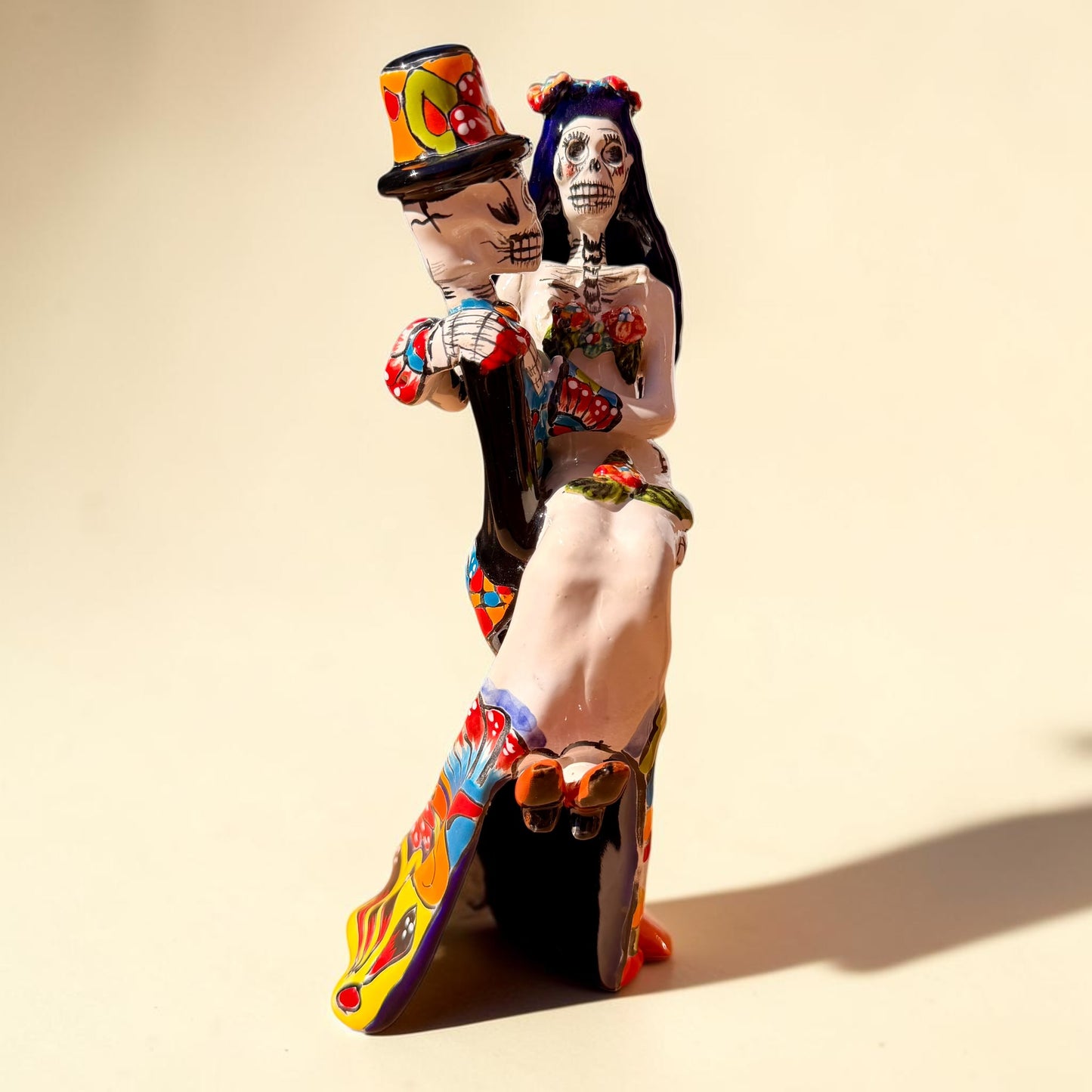 A colorful Day of the Dead figurine of a skeleton bride and groom embracing, with the groom holding the bride. The figurine features vibrant painted details including a orange and white patterned dress, a multicolored hat, and dark hair, set against a plain light background. Side view showing brides feet.