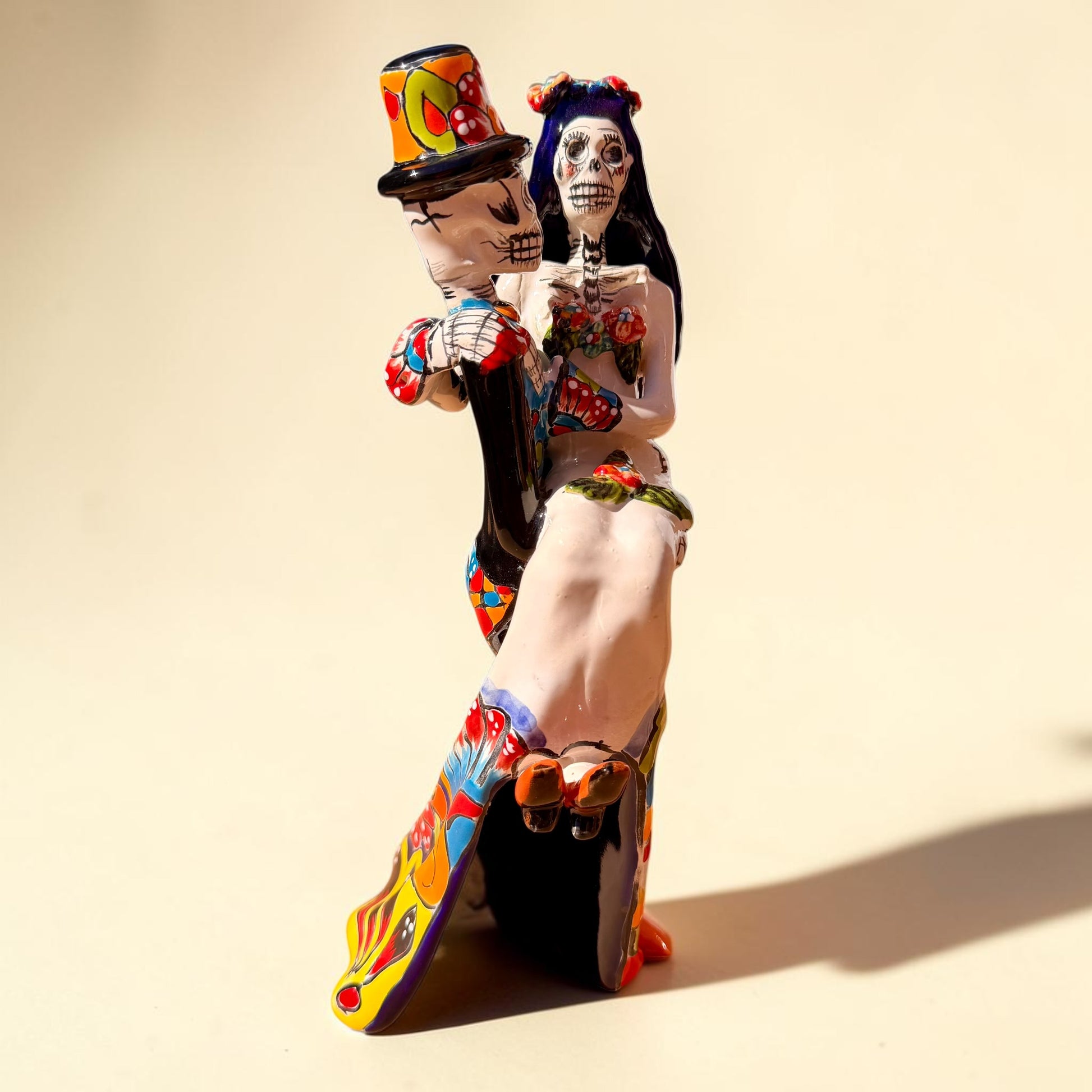 A colorful Day of the Dead figurine of a skeleton bride and groom embracing, with the groom holding the bride. The figurine features vibrant painted details including a orange and white patterned dress, a multicolored hat, and dark hair, set against a plain light background. Side view showing brides feet.