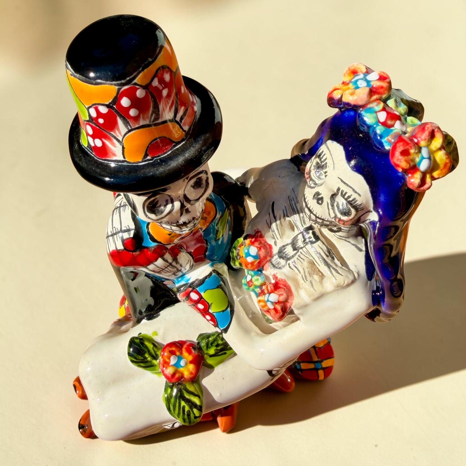 A colorful Day of the Dead figurine of a skeleton bride and groom embracing, with the groom holding the bride. The figurine features vibrant painted details including a orange and white patterned dress, a multicolored hat, and dark hair, set against a plain light background. Top view of couple.