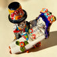 A colorful Day of the Dead figurine of a skeleton bride and groom embracing, with the groom holding the bride. The figurine features vibrant painted details including a orange and white patterned dress, a multicolored hat, and dark hair, set against a plain light background. Top view of couple.