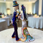 Mexican Talavera Catrina Bride and Groom dancing at their wedding.  He is wearing a tuxcedo with a top hat and she is wearing a flowing wedding dress with a veil and floral pattern toward the bottom.  They are at their wedding reception.