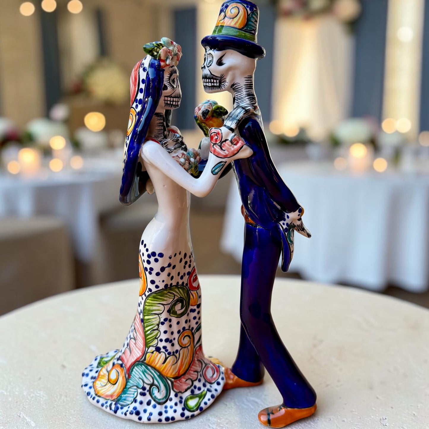 Mexican Talavera Catrina Bride and Groom dancing at their wedding.  He is wearing a tuxcedo with a top hat and she is wearing a flowing wedding dress with a veil and floral pattern toward the bottom.  Opposite side view of couple.