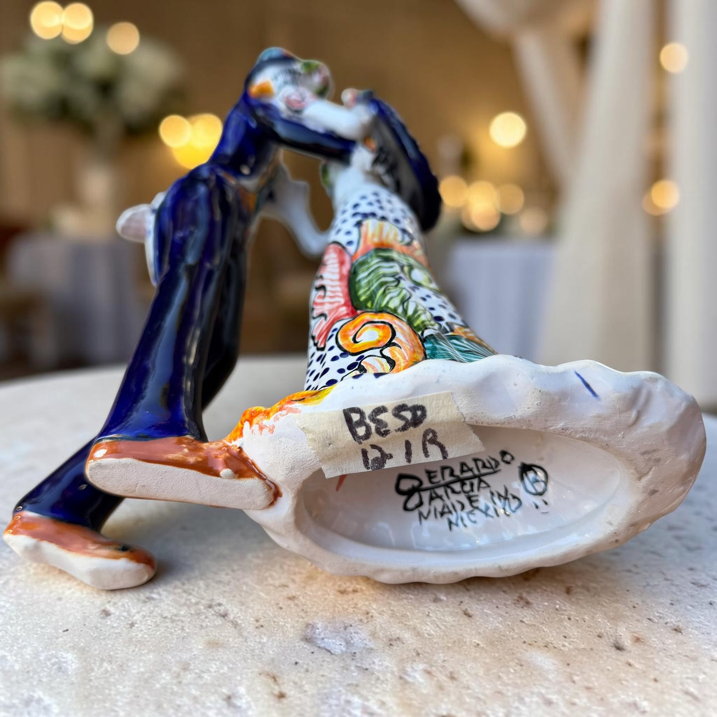 Mexican Talavera Catrina Bride and Groom dancing at their wedding.  He is wearing a tuxcedo with a top hat and she is wearing a flowing wedding dress with a veil and floral pattern toward the bottom.  They are at their wedding reception. Bottom view showing artist signature.