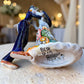 Mexican Talavera Catrina Bride and Groom dancing at their wedding.  He is wearing a tuxcedo with a top hat and she is wearing a flowing wedding dress with a veil and floral pattern toward the bottom.  They are at their wedding reception. Bottom view showing artist signature.