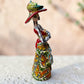 Mexican catrina talavera doll with calla lilies on the front of her dress. wearing a colorful dress with a big feathered hat.   Left side view of doll.