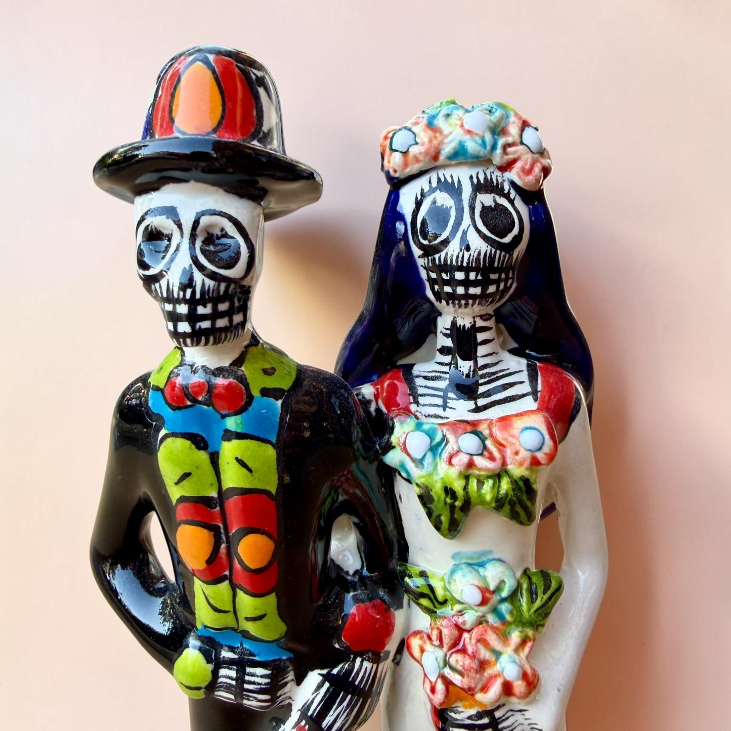 A talavera Day of the Dead wedding couple figurine featuring a groom skeleton in a black suit with colorful floral patterns and a bride skeleton in a white wedding gown with floral details, set against a solid pale peach background. Close up of the faces.