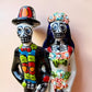 A talavera Day of the Dead wedding couple figurine featuring a groom skeleton in a black suit with colorful floral patterns and a bride skeleton in a white wedding gown with floral details, set against a solid pale peach background. Close up of the faces.