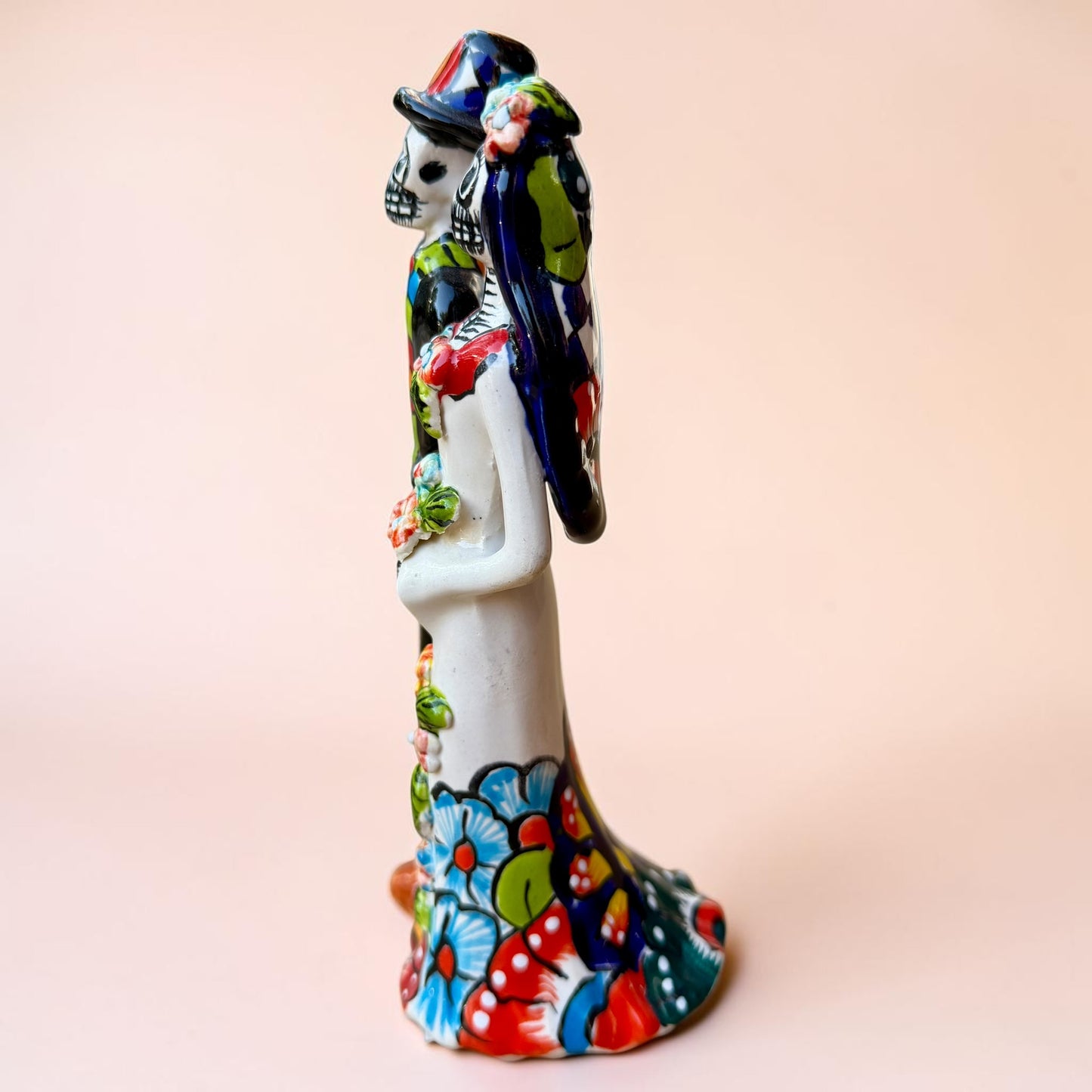 A talavera Day of the Dead wedding couple figurine featuring a groom skeleton in a black suit with colorful floral patterns and a bride skeleton in a white wedding gown with floral details, set against a solid pale peach background. Right side view.
