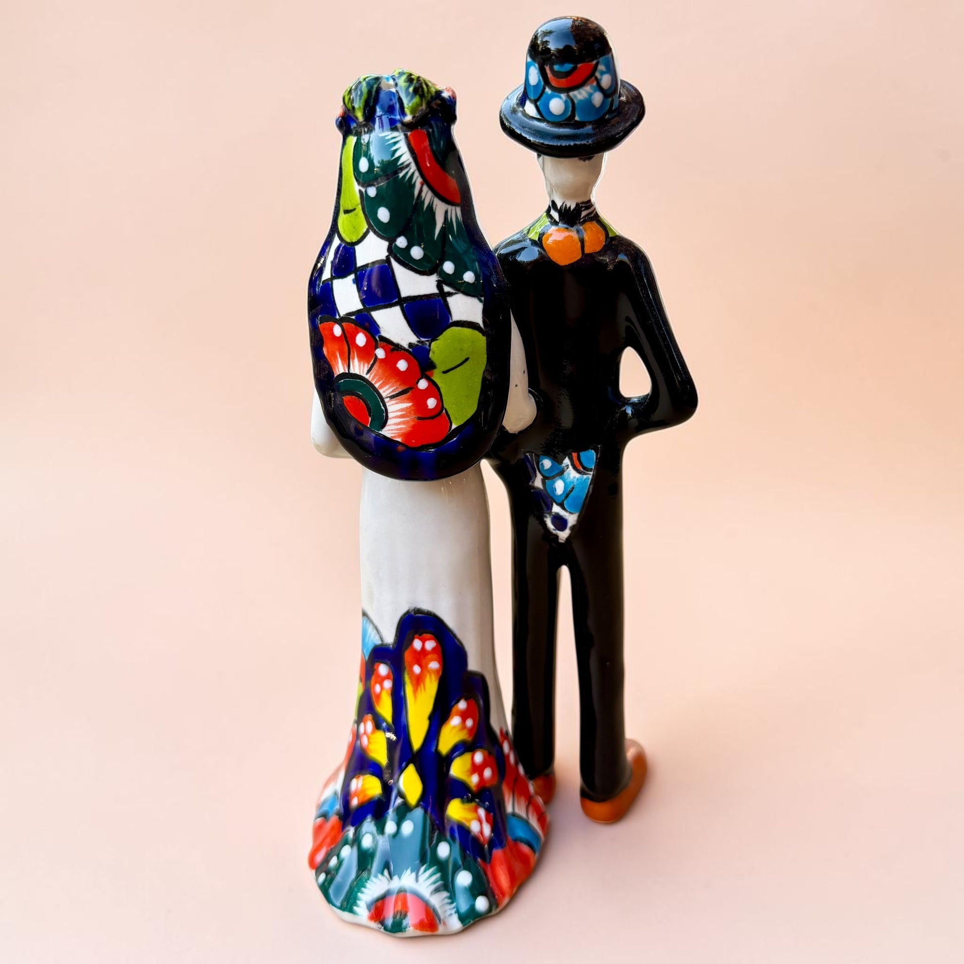 A talavera Day of the Dead wedding couple figurine featuring a groom skeleton in a black suit with colorful floral patterns and a bride skeleton in a white wedding gown with floral details, set against a solid pale peach background. Back side view of couple showing veil and dress train.