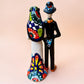 A talavera Day of the Dead wedding couple figurine featuring a groom skeleton in a black suit with colorful floral patterns and a bride skeleton in a white wedding gown with floral details, set against a solid pale peach background. Back side view of couple showing veil and dress train.
