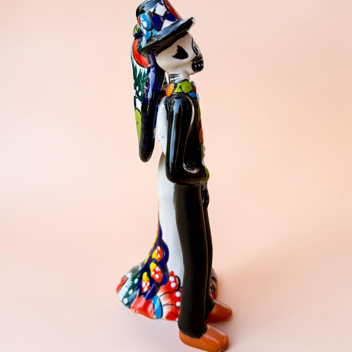 A talavera Day of the Dead wedding couple figurine featuring a groom skeleton in a black suit with colorful floral patterns and a bride skeleton in a white wedding gown with floral details, set against a solid pale peach background. Left side view focusing on the groom.