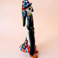 A talavera Day of the Dead wedding couple figurine featuring a groom skeleton in a black suit with colorful floral patterns and a bride skeleton in a white wedding gown with floral details, set against a solid pale peach background. Left side view focusing on the groom.