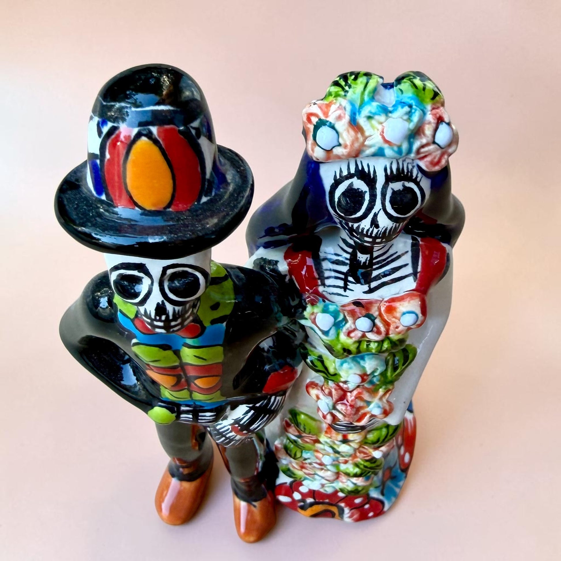 A talavera Day of the Dead wedding couple figurine featuring a groom skeleton in a black suit with colorful floral patterns and a bride skeleton in a white wedding gown with floral details, set against a solid pale peach background. Top view of couple.