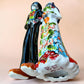 A talavera Day of the Dead wedding couple figurine featuring a groom skeleton in a black suit with colorful floral patterns and a bride skeleton in a white wedding gown with floral details, set against a solid pale peach background.  Bottom View.