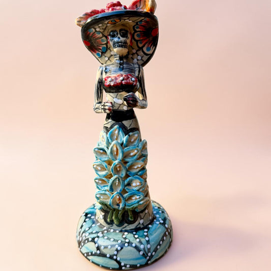 A hand-painted Day of the Dead (Día de los Muertos) talavera figurine of a skeleton woman featuring calla lily her her dress and wearing a large floral sombrero, set against a solid pale pink background.