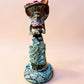 A hand-painted Day of the Dead (Día de los Muertos) talavera figurine of a skeleton woman featuring calla lily her her dress and wearing a large floral sombrero, set against a solid pale pink background.