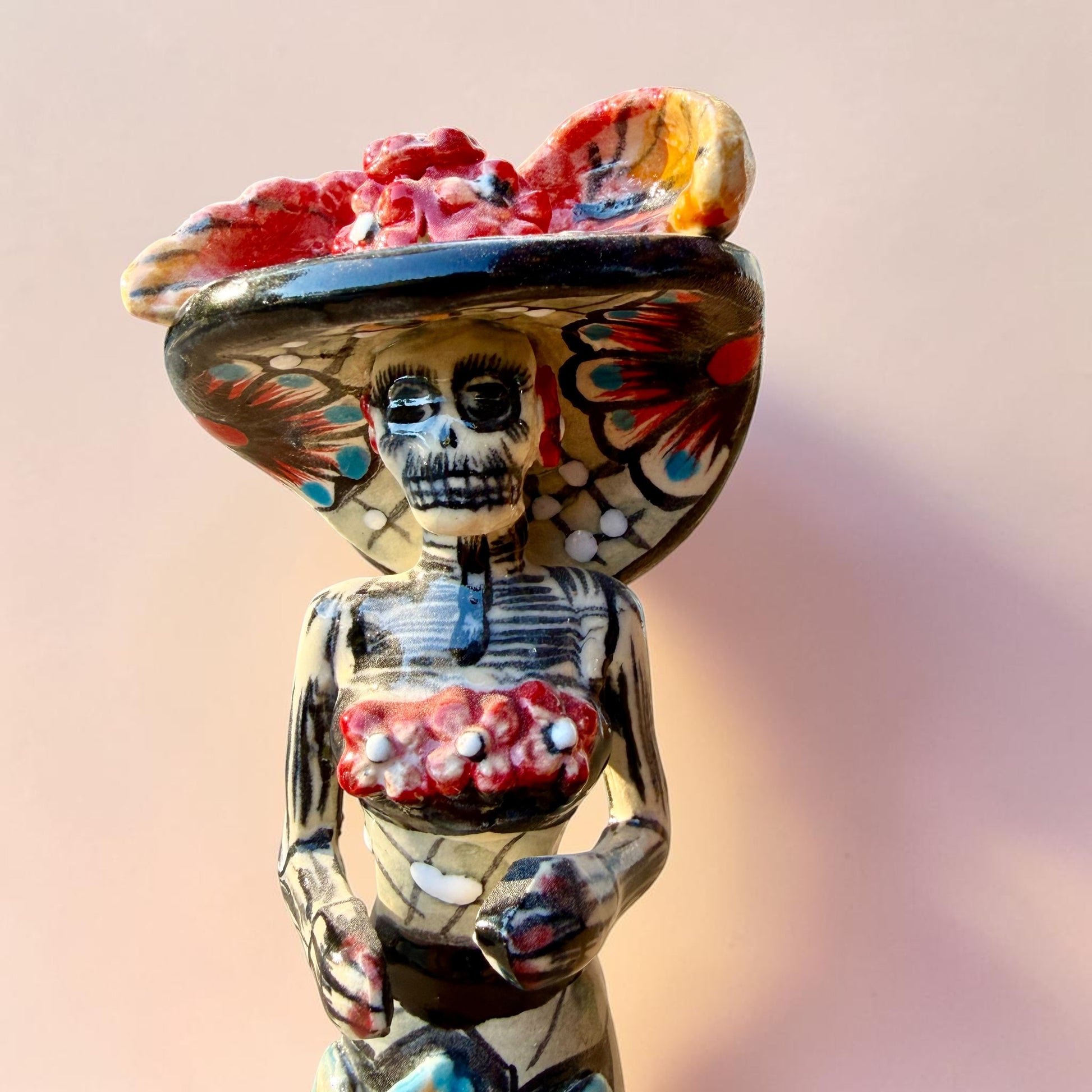 A hand-painted Day of the Dead (Día de los Muertos) talavera figurine of a skeleton woman featuring calla lily her her dress and wearing a large floral sombrero, set against a solid pale pink background. Face close up of the doll.