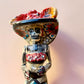 A hand-painted Day of the Dead (Día de los Muertos) talavera figurine of a skeleton woman featuring calla lily her her dress and wearing a large floral sombrero, set against a solid pale pink background. Face close up of the doll.