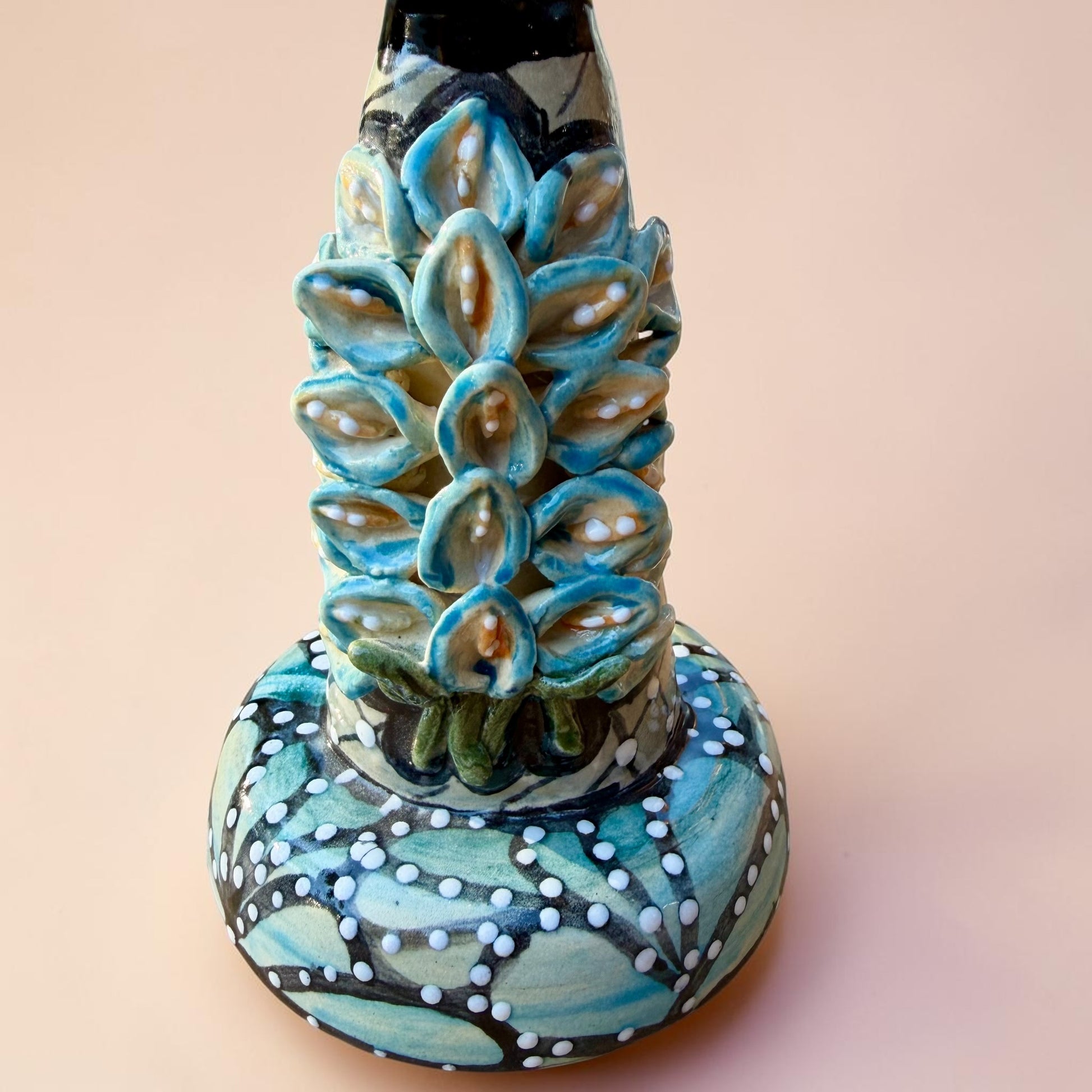 A hand-painted Day of the Dead (Día de los Muertos) talavera figurine of a skeleton woman featuring calla lily her her dress and wearing a large floral sombrero, set against a solid pale pink background. Close up of the blue tinted Calla Lilies.