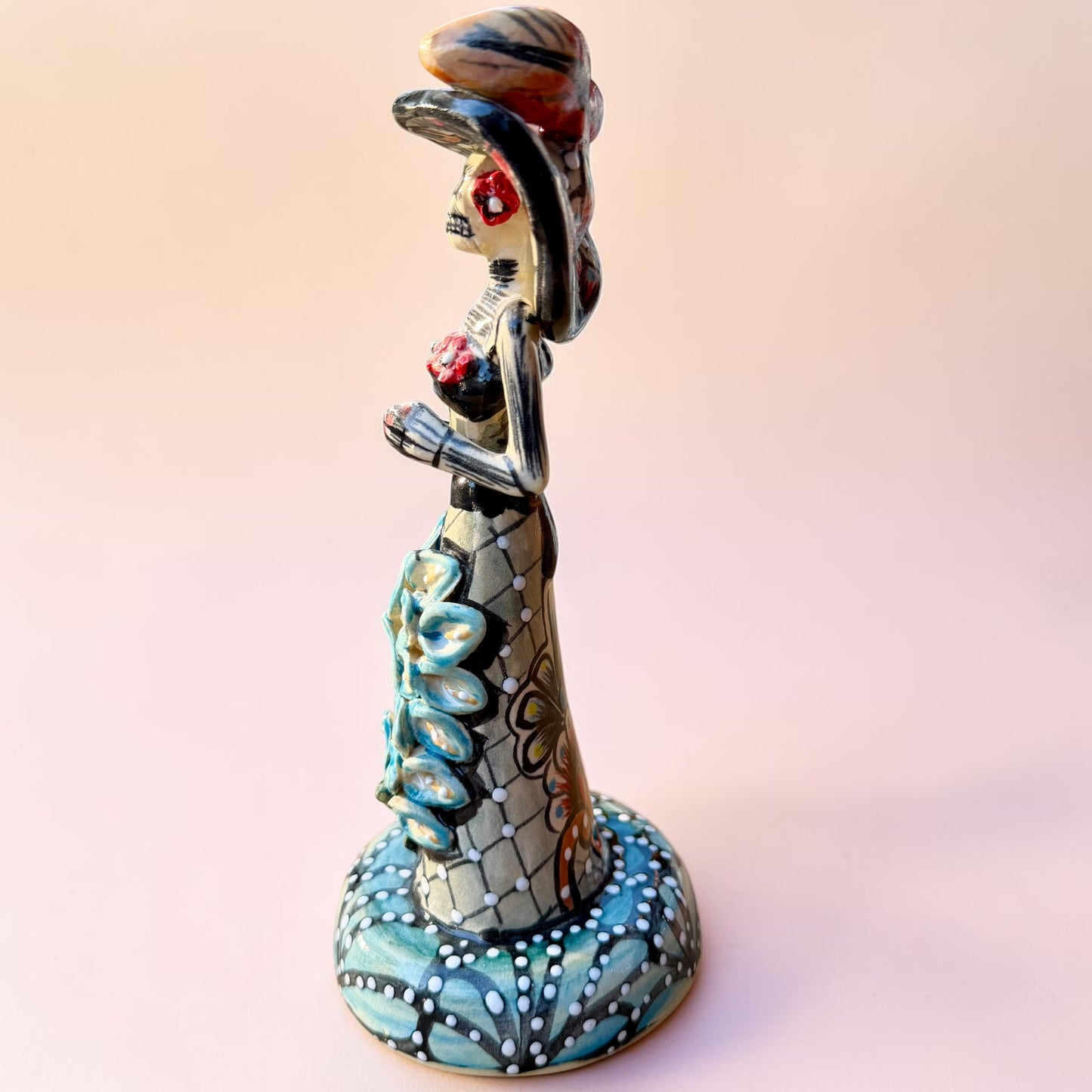 A hand-painted Day of the Dead (Día de los Muertos) talavera figurine of a skeleton woman featuring calla lily her her dress and wearing a large floral sombrero, set against a solid pale pink background. Right side view of doll.