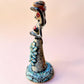A hand-painted Day of the Dead (Día de los Muertos) talavera figurine of a skeleton woman featuring calla lily her her dress and wearing a large floral sombrero, set against a solid pale pink background. Right side view of doll.