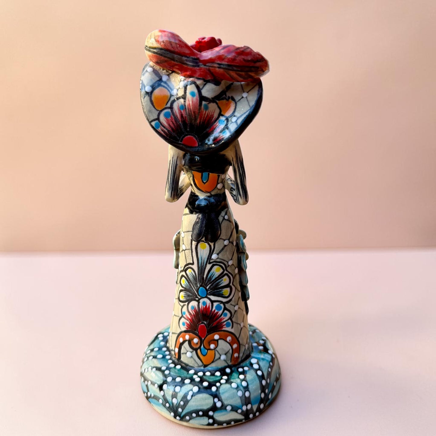 A hand-painted Day of the Dead (Día de los Muertos) talavera figurine of a skeleton woman featuring calla lily her her dress and wearing a large floral sombrero, set against a solid pale pink background. Back view of catrina doll.