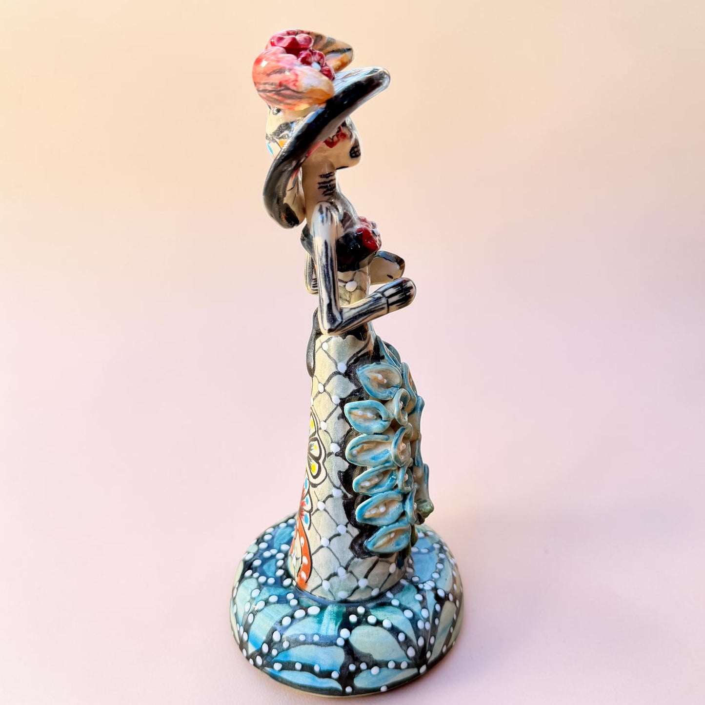 A hand-painted Day of the Dead (Día de los Muertos) talavera figurine of a skeleton woman featuring calla lily her her dress and wearing a large floral sombrero, set against a solid pale pink background. Left side view of the catrina doll.