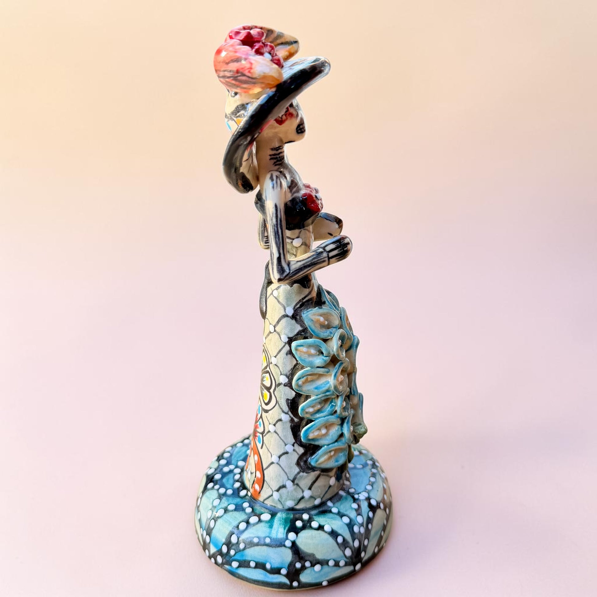 A hand-painted Day of the Dead (Día de los Muertos) talavera figurine of a skeleton woman featuring calla lily her her dress and wearing a large floral sombrero, set against a solid pale pink background. Left side view of the catrina doll.