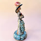 A hand-painted Day of the Dead (Día de los Muertos) talavera figurine of a skeleton woman featuring calla lily her her dress and wearing a large floral sombrero, set against a solid pale pink background. Left side view of the catrina doll.