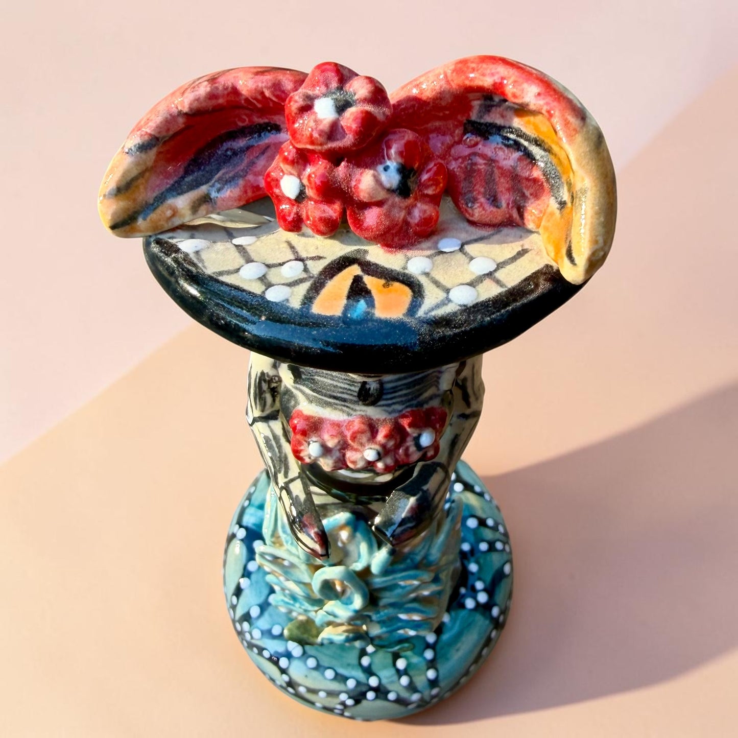 A hand-painted Day of the Dead (Día de los Muertos) talavera figurine of a skeleton woman featuring calla lily her her dress and wearing a large floral sombrero, set against a solid pale pink background. Top view of the catrina doll showing feather decorated hat.
