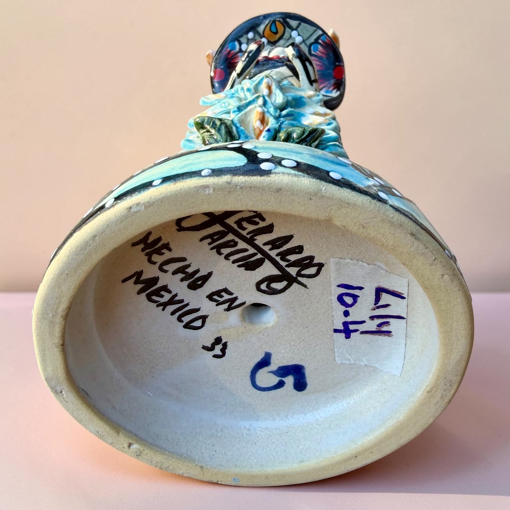 A hand-painted Day of the Dead (Día de los Muertos) talavera figurine of a skeleton woman featuring calla lily her her dress and wearing a large floral sombrero, set against a solid pale pink background. Bottom showing artist signature.