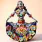 A colorful hand-painted Day of the Dead (Día de los Muertos) ceramic figurine of a  ballerina, decorated with vibrant floral patterns and a white skull head with braided hair, set against a solid peach background. Front view of entire figurine.