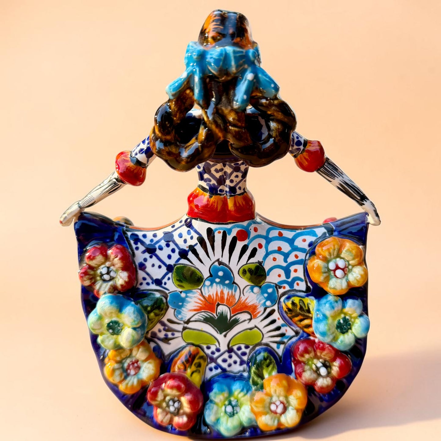 A colorful hand-painted Day of the Dead (Día de los Muertos) ceramic figurine of a  ballerina, decorated with vibrant floral patterns and a white skull head with braided hair, set against a solid peach background. Back view of the ballerina doll showing her braided pig tails.