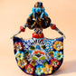 A colorful hand-painted Day of the Dead (Día de los Muertos) ceramic figurine of a  ballerina, decorated with vibrant floral patterns and a white skull head with braided hair, set against a solid peach background. Back view of the ballerina doll showing her braided pig tails.